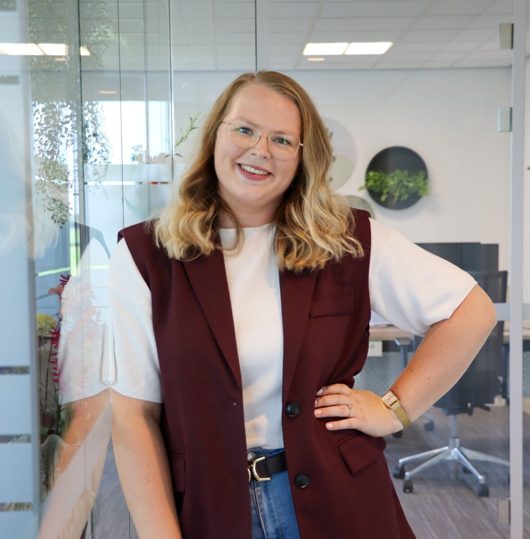Smiling woman wearing a white top and maroon vest stands with hand on hip inside a glass-walled office. She wears glasses and jeans.