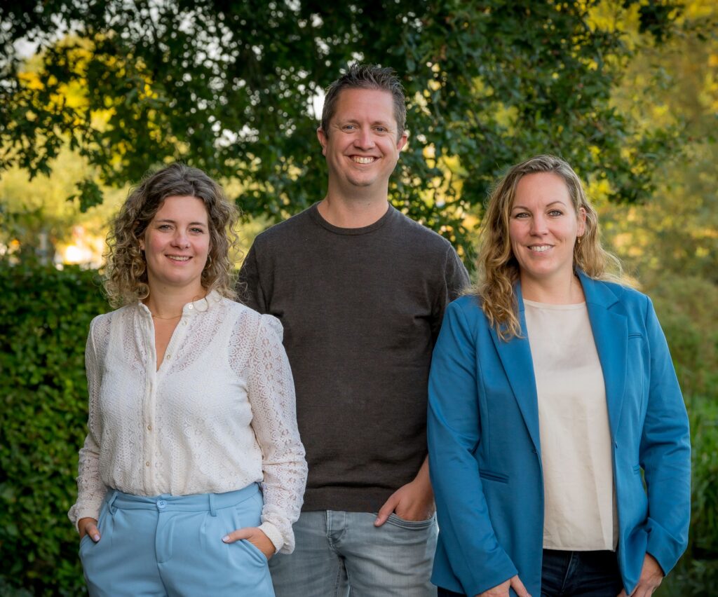 Three adults smiling outdoors, left woman in a white lace blouse, center man in a dark T‑shirt, right woman in a blue blazer with a beige top.