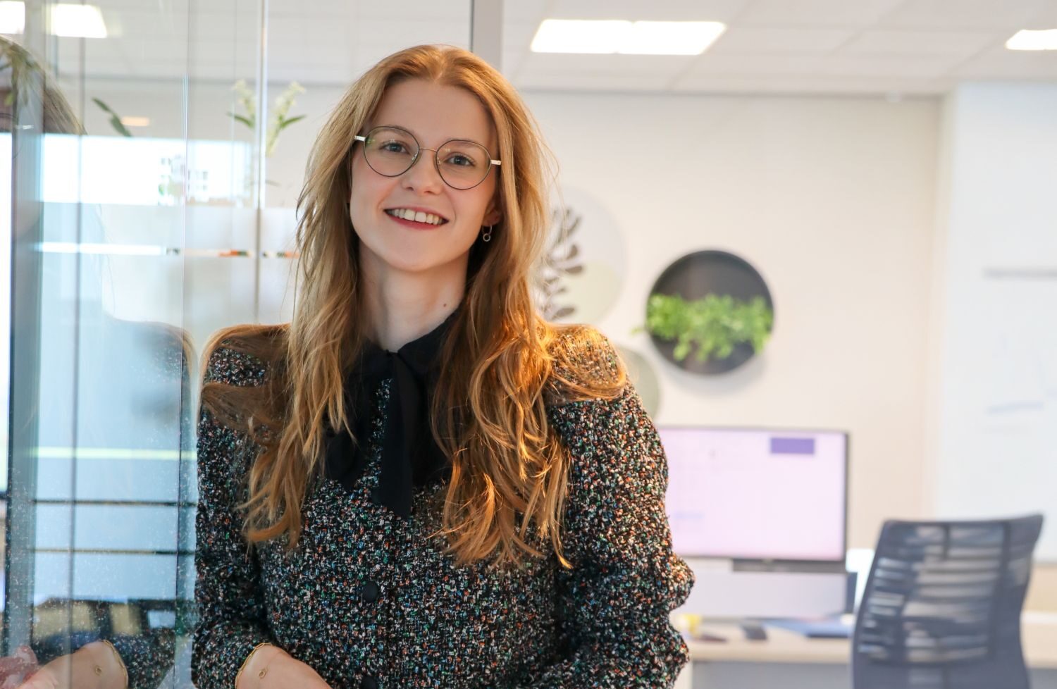 Smiling woman with round glasses and long blond hair leaning on a glass wall in a bright office, wearing a dark patterned blazer with a black bow tie.