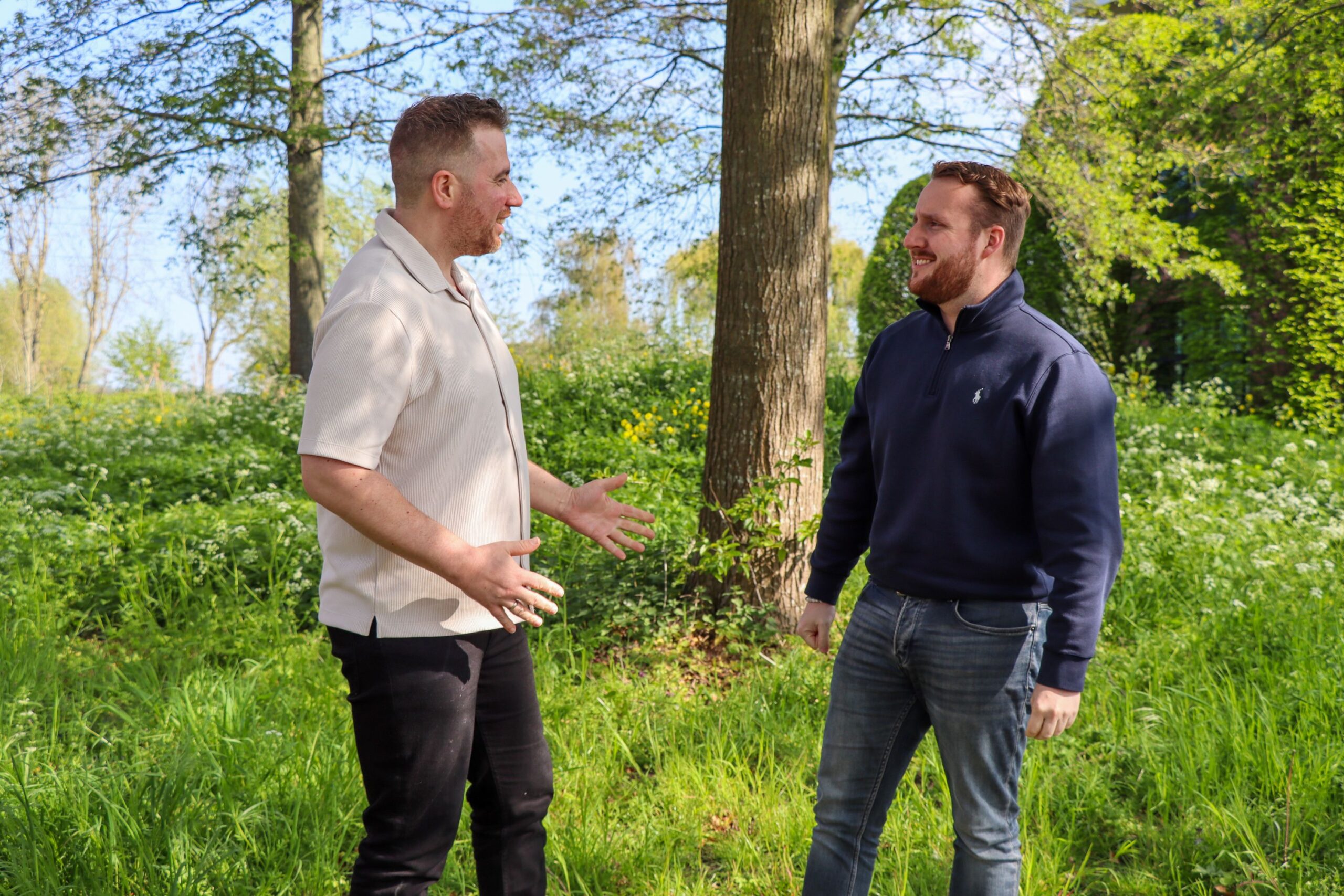 Two men stand in a sunny park, facing and talking among tall trees and green plants in bloom.