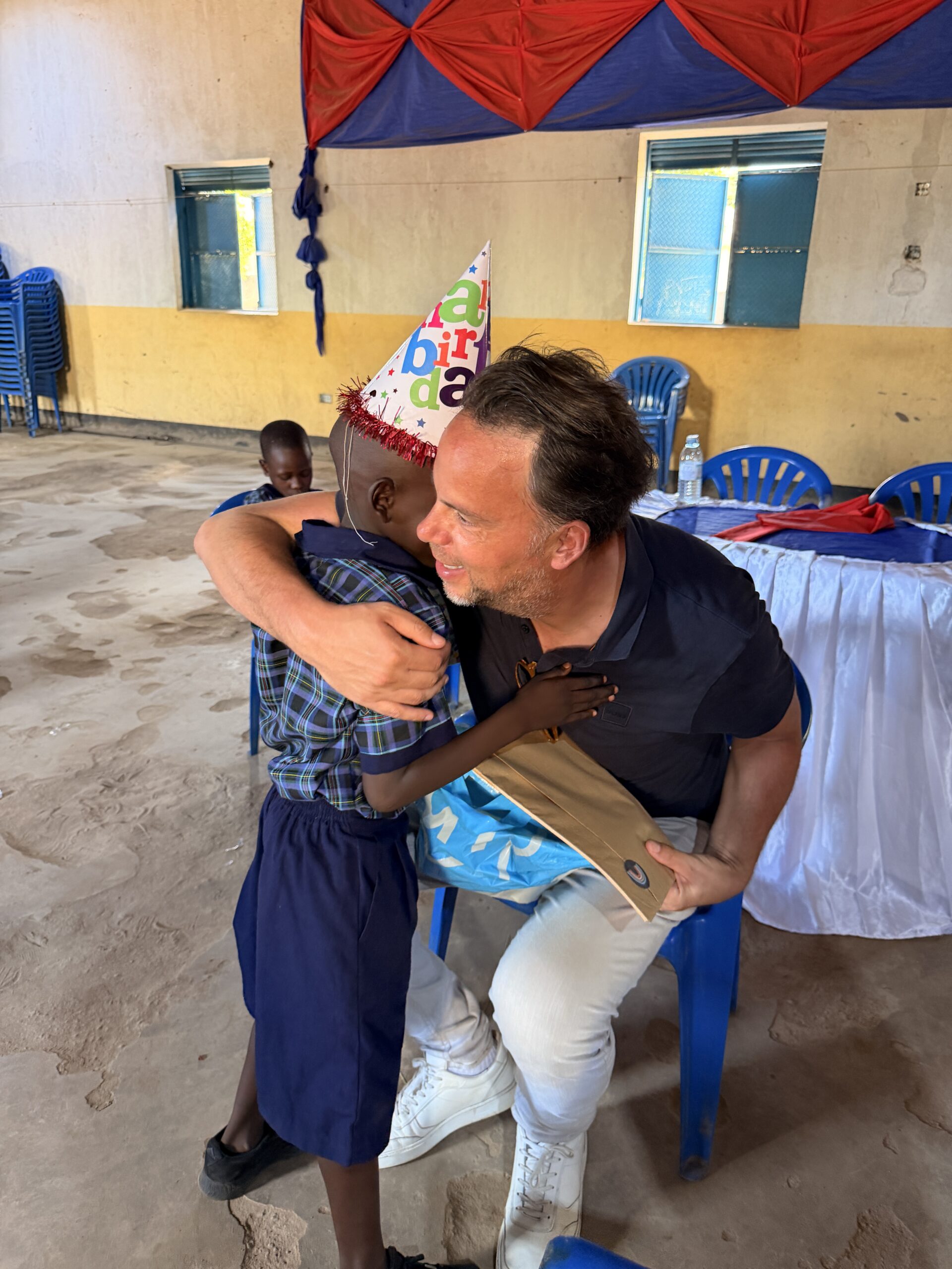 Man and young boy share a warm hug at a birthday party; boy wears a colorful 'birthday' hat in a hall with blue chairs and red-blue decorations.