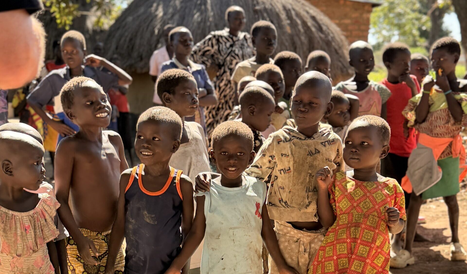 Group of children standing together outdoors in a sunny dusty yard, some barefoot, colorful clothing, with a thatched hut in the background.