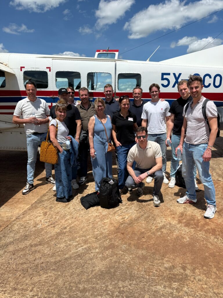 Group of people posing for a photo in front of a small white airplane with red/blue stripes behind them, outdoors on a sunny day.