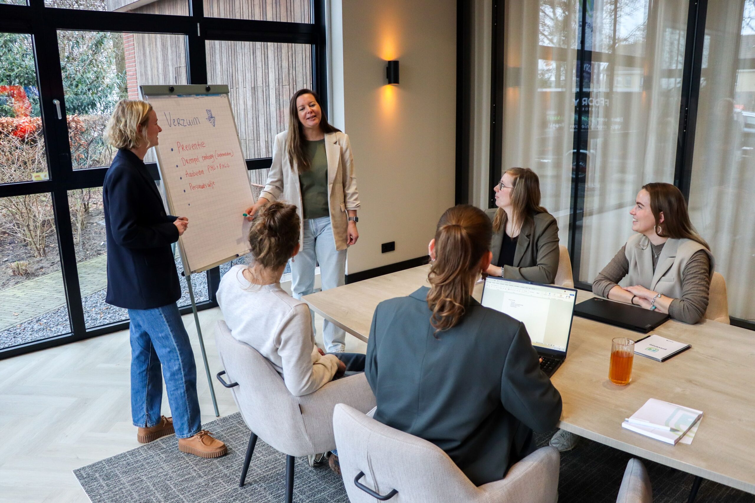 Presenter standing by flip chart briefing four colleagues in a glass-walled meeting room.