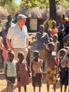 A man in a light shirt and flat cap stands with a group of barefoot children in a rural village with thatched huts in the background, engaging with them.