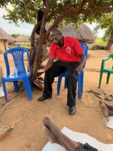 Man in a red polo sits on a blue plastic chair under a tree in a rural village, smiling as he gestures with his hand; huts and barefoot people in the background.