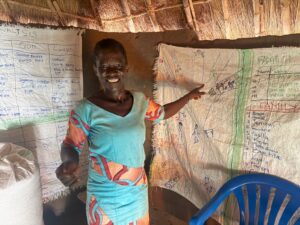 Smiling woman standing beside large fabric charts with handwritten notes and drawings taped to the wall in a thatched room.