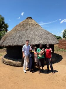 Man in white standing with local family in front of a round thatched hut on a sunny day, group smiling outside a village home.