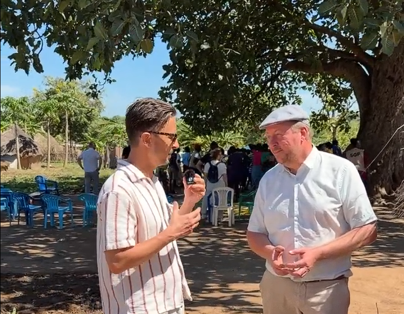 Two men conversing outdoors under a large tree at a sunny gathering, with blue plastic chairs in the background.