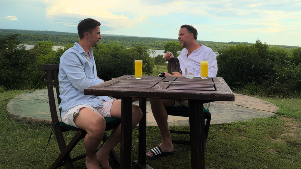 Two men sit at a weathered wooden table outdoors, chatting, with two yellow juice drinks and a water glass against a green landscape.