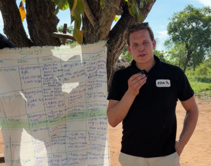 Man in a black polo with a ZoA logo stands outdoors beside a large handwritten chart, explaining its content to the camera, under a tree.
